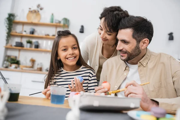 Smiling kid looking at camera near parents while coloring Easter eggs at home — Stock Photo