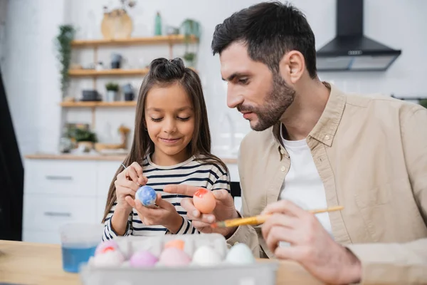 Smiling girl painting Easter eggs with father in kitchen — Photo de stock