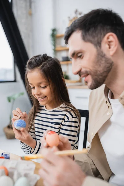 Smiling girl painting Easter eggs near blurred father at home — Photo de stock