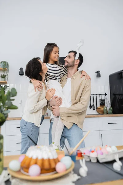 Pais segurando filha perto de bolo de Páscoa borrado e ovos em casa — Fotografia de Stock