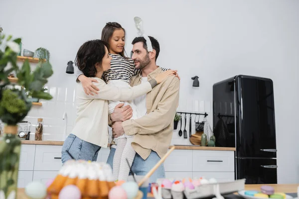 Positive parents hugging daughter near blurred Easter eggs and cake in kitchen — Stock Photo