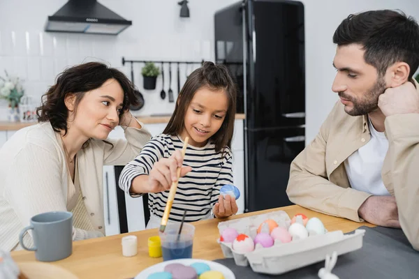 Pais olhando para a filha feliz colorir ovos de Páscoa perto macaroons em casa — Fotografia de Stock