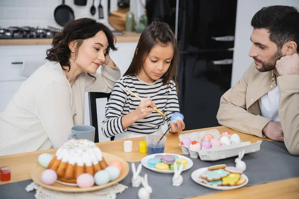 Família sentada perto da filha colorir ovos de Páscoa perto de comida em casa — Fotografia de Stock