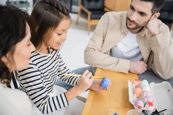 Smiling girl painting Easter egg near mother and father at home — Photo de stock