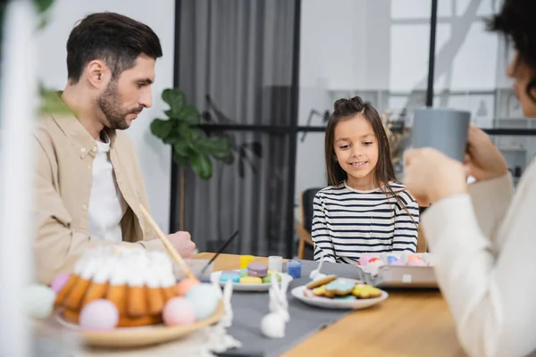 Smiling girl sitting near Easter eggs and cake beside parents at home — Stock Photo