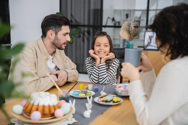 Criança alegre sentada perto dos pais e comida de Páscoa saborosa em casa — Fotografia de Stock