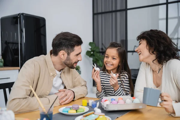 Smiling parents looking at happy daughter with Easter rabbits near eggs and macaroons at home — Stock Photo