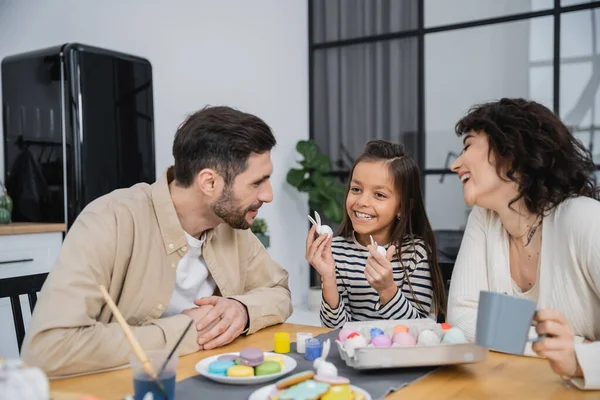 Smiling parents looking at happy daughter with Easter rabbits near eggs and macaroons at home — Photo de stock