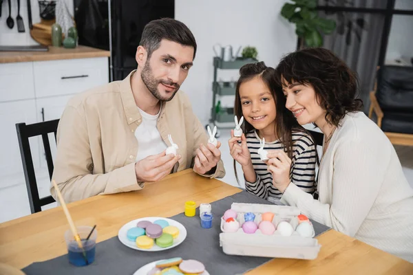 Pais e criança segurando decoração de Páscoa perto de ovos e macaroons na cozinha — Fotografia de Stock