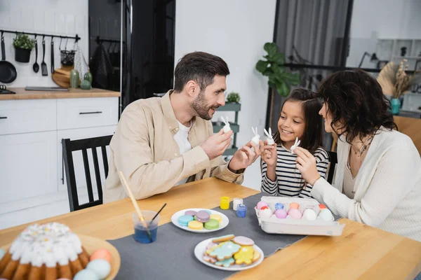 Positive parents and kid holding Easter rabbits near cake and eggs at home — Fotografia de Stock