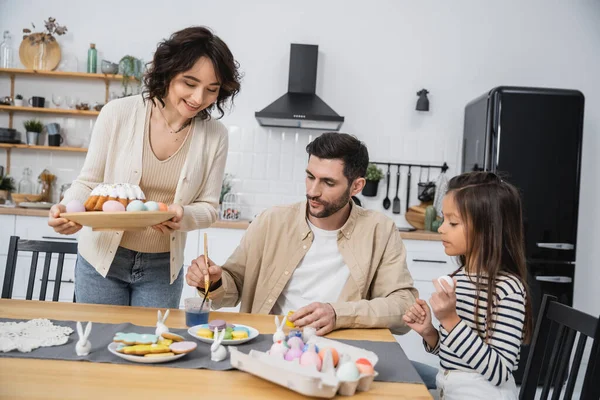 Mulher positiva segurando prato com bolo de Páscoa enquanto a família colorir ovos em casa — Fotografia de Stock