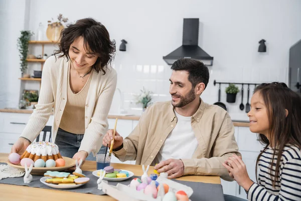 Family coloring Easter eggs near happy woman with cake in kitchen — Stock Photo