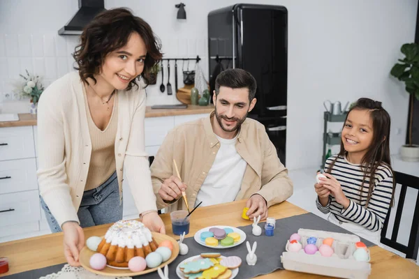Familia para colorear huevos de Pascua cerca de la madre alegre celebración plato con pastel en casa - foto de stock