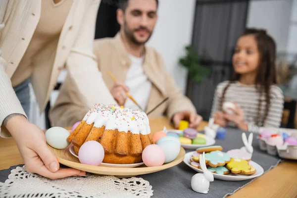 Woman putting plate with Easter cake on table near blurred family at home — Stock Photo