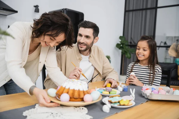 Sorrindo homem e menina colorir ovos de Páscoa perto de mulher com bolo em casa — Fotografia de Stock