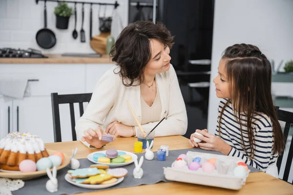 Усміхнена дівчина розмовляє з мамою біля великодніх яєць і солодкої їжі на столі — стокове фото