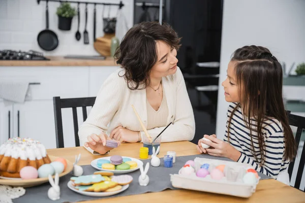 Smiling girl talking to mom near Easter eggs and sweet food on table — Stock Photo