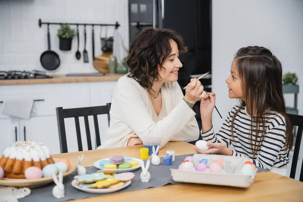 Frau und Tochter halten Pinsel neben bunten Ostereiern und Kuchen zu Hause — Stockfoto