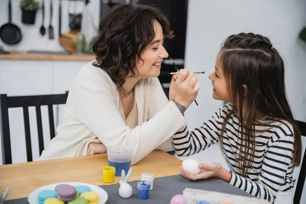 Side view of cheerful mother and child holding paintbrushes near Easter eggs and macaroons at home — Stock Photo