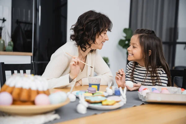 Positive mother and kid holding paintbrushes while coloring Easter eggs at home — Stock Photo
