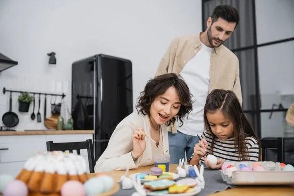 Excited mother and daughter coloring Easter eggs near man in kitchen — Stock Photo