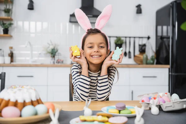 Весела дитина в хустці тримає печиво біля великодніх торта та яєць вдома — стокове фото