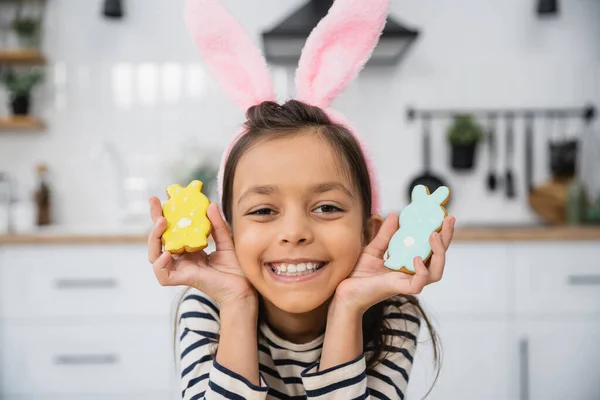 Smiling kid in Easter headband with bunny ears holding cookies in kitchen — Stock Photo