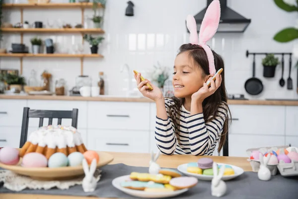 Smiling child in headband looking at Easter cookie near food in kitchen — Fotografia de Stock