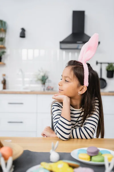 Preteen kid in bunny headband sitting near macaroons and Easter decor at home — Fotografia de Stock