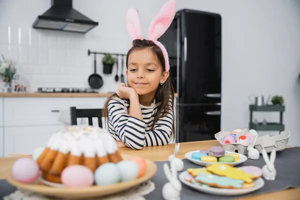 Preteen kid in bunny ears headband looking at Easter cake and eggs in kitchen — Fotografia de Stock