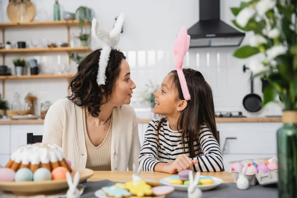 Vista lateral de mãe sorridente e criança em headbands sentado perto de comida de Páscoa em casa — Fotografia de Stock