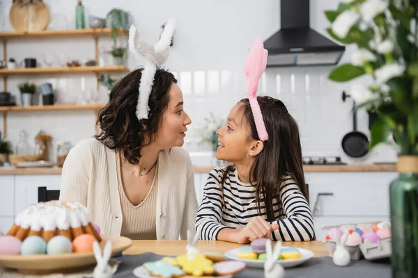 Side view of smiling mom and kid in headbands sitting near Easter food at home — Stock Photo