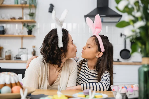Side view of mom and daughter in headbands pouting lips near Easter food in kitchen — Fotografia de Stock