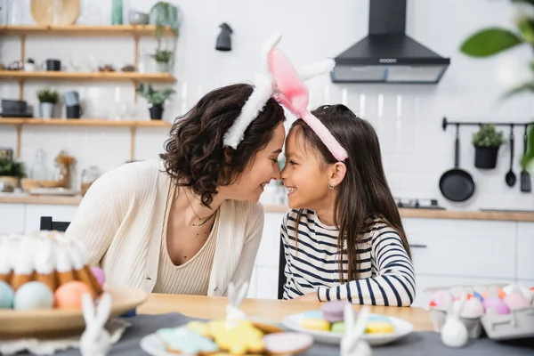 Vista lateral de mãe e filha sorridente na Páscoa headbands sentado perto de comida na cozinha — Fotografia de Stock