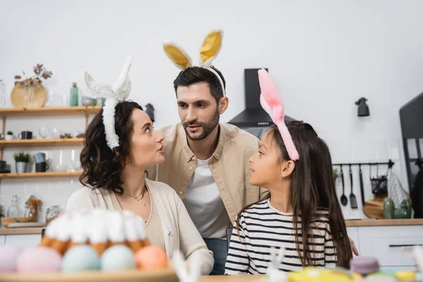 Family in bunny ears headbands sitting near blurred Easter Cake in kitchen — Fotografia de Stock