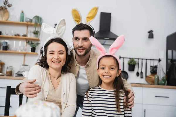 Smiling man hugging wife and daughter in Easter headbands in kitchen — Fotografia de Stock