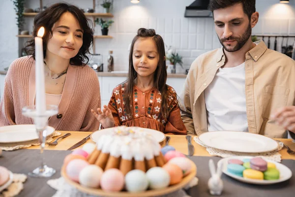 Family sitting near blurred Easter cake and candle on table at home — Fotografia de Stock