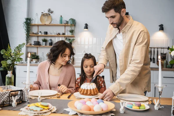 Pai cortando bolo de Páscoa perto de filha e esposa durante o jantar festivo — Fotografia de Stock
