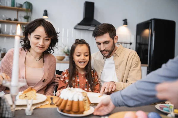 Familia alegre sentada cerca de huevos de Pascua y pastel durante la cena en casa - foto de stock