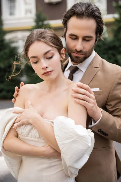 Бородатий наречений в офіційному одязі обіймає гарну наречену в білій сукні — стокове фото