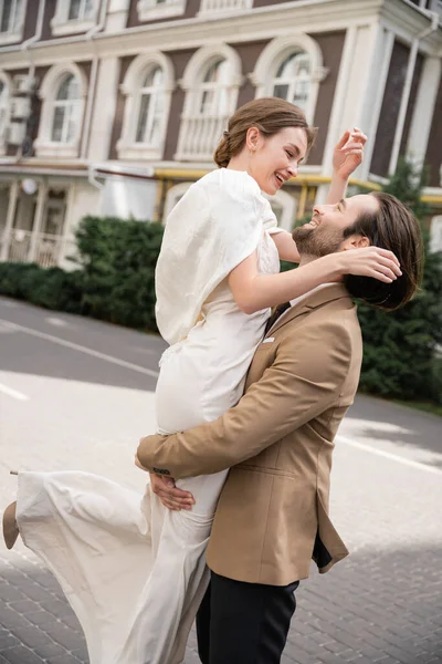 Bearded groom in formal wear lifting happy bride in white dress outside — Stock Photo