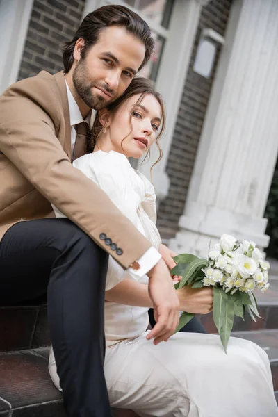 Bearded groom and pretty bride with wedding bouquet looking at camera — Stockfoto