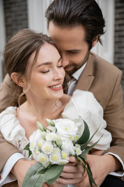 Bärtiger Bräutigam umarmt glückliche Braut im weißen Kleid mit Brautstrauß — Stockfoto