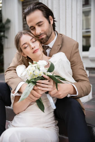Bearded groom with closed eyes hugging gorgeous bride in white dress with wedding bouquet — Stock Photo