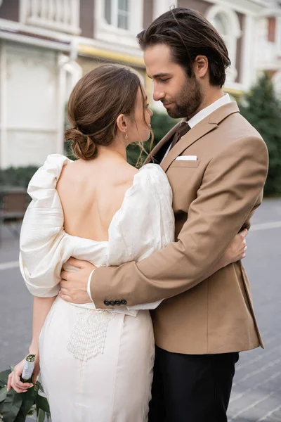 Noivo barbudo em terno abraçando noiva em vestido branco com buquê de casamento — Fotografia de Stock