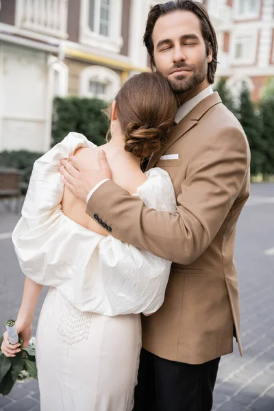Novio elegante y barbudo con los ojos cerrados abrazando novia en vestido blanco - foto de stock