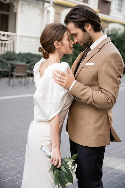 Side view of bearded groom in suit hugging bride in white dress with wedding bouquet — Foto stock