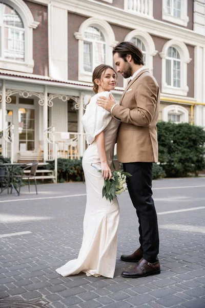 Повна довжина бородатого нареченого в костюмі обіймає щасливу наречену в білій сукні з весільним букетом, стоячи на вулиці — стокове фото
