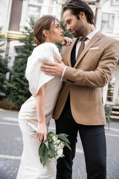 Bearded groom in suit hugging young bride in white dress with wedding bouquet while standing on street — Photo de stock
