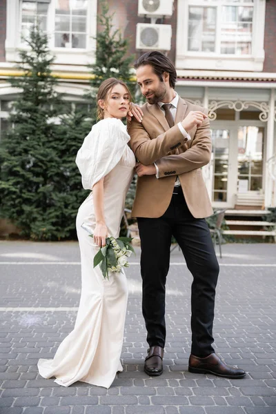 Повна довжина молодої нареченої в білій сукні, що тримає весільний букет, стоячи біля веселого нареченого в костюмі — стокове фото